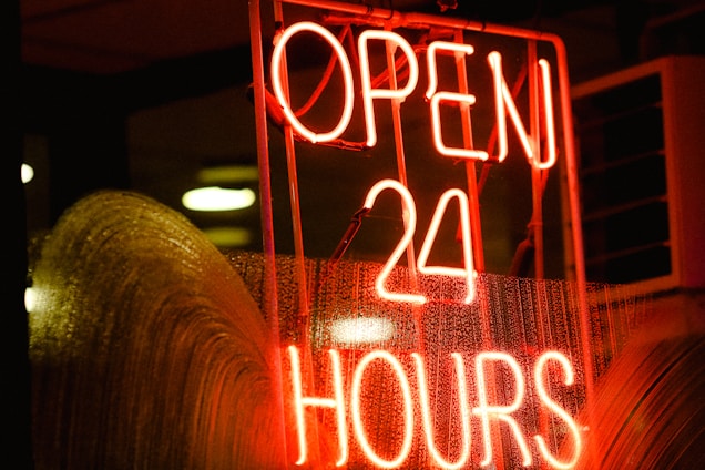 A bright neon sign with the words 'OPEN 24 HOURS' in bold red letters. The sign is lit up against a dark background, with droplets of water visible on the glass surface in front of the sign, likely due to rain.