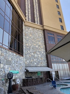 A tall building with a stone facade and large windows dominates the scene. In the foreground, there are poolside loungers, a standing fan, and a small swimming pool with a pool lift chair. An awning extends over a bar area with high chairs, and a sign about pool regulations is visible.