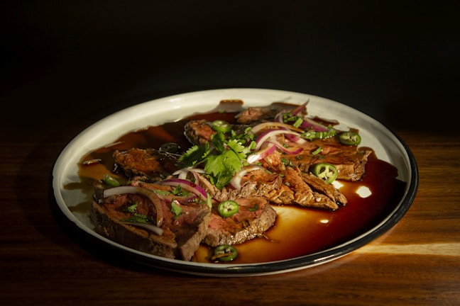 Slices of tender grilled beef are arranged on a white plate, drizzled with a rich brown sauce. The dish is garnished with fresh green jalapeño slices, red onion rings, and cilantro, adding vibrant color and freshness.