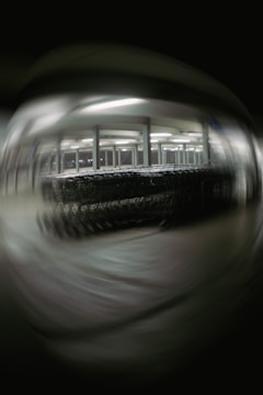 A blurred image depicting a large number of shopping carts lined up under bright fluorescent lights in a structured setting, possibly a warehouse or a storage area. The background and foreground are out of focus, creating a distorted effect.