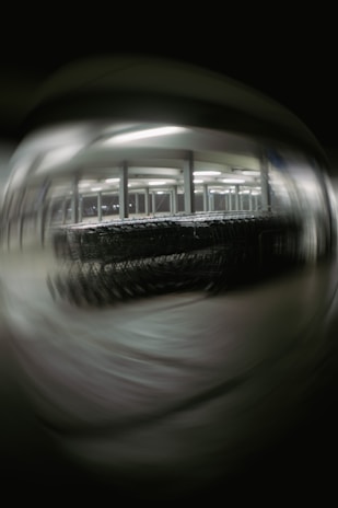 A blurred image depicting a large number of shopping carts lined up under bright fluorescent lights in a structured setting, possibly a warehouse or a storage area. The background and foreground are out of focus, creating a distorted effect.