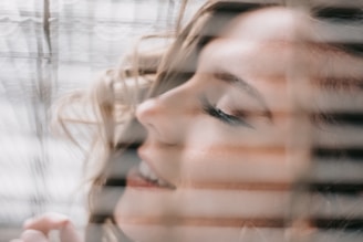 A serene and softly focused close-up of a woman's face with her eyes gently closed. Her expression suggests a sense of relaxation and contentment. Her face is framed by soft, wavy hair, and seen through what appears to be window blinds, creating lines of shadow and light across her features.