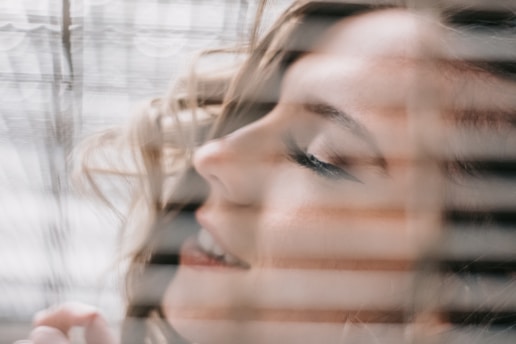 A serene and softly focused close-up of a woman's face with her eyes gently closed. Her expression suggests a sense of relaxation and contentment. Her face is framed by soft, wavy hair, and seen through what appears to be window blinds, creating lines of shadow and light across her features.