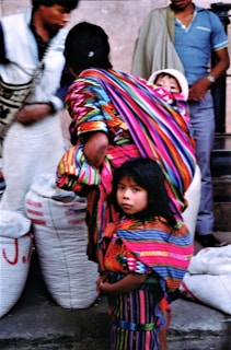 A young girl with long dark hair is wearing a vibrantly colored traditional garment with stripes of pink, blue, orange, and green. She stands among a small crowd, next to another person bundled in a similar colorful cloth. A baby peeks out from the folds of the fabric the adult is wearing. Large sacks are visible in the background, suggesting a market or outdoor setting.