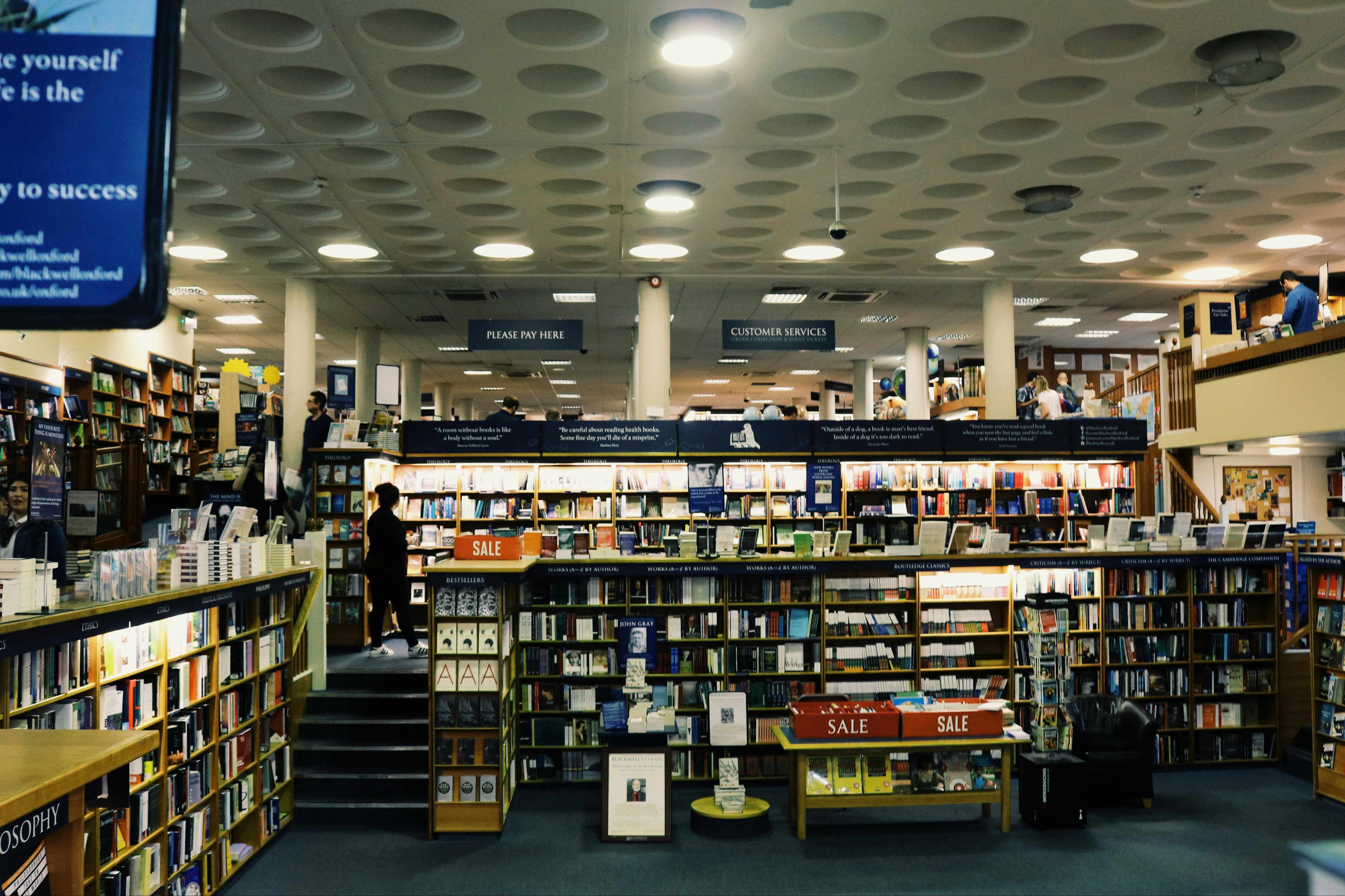 A spacious bookstore with multiple shelves filled with books, including a sale section in the center. The store features a modern design with a high ceiling and circular light fixtures. Several people are browsing the books, and signs such as 'PLEASE PAY HERE' and 'CUSTOMER SERVICES' are visible.