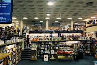 A spacious bookstore with multiple shelves filled with books, including a sale section in the center. The store features a modern design with a high ceiling and circular light fixtures. Several people are browsing the books, and signs such as 'PLEASE PAY HERE' and 'CUSTOMER SERVICES' are visible.