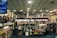 A spacious bookstore with multiple shelves filled with books, including a sale section in the center. The store features a modern design with a high ceiling and circular light fixtures. Several people are browsing the books, and signs such as 'PLEASE PAY HERE' and 'CUSTOMER SERVICES' are visible.