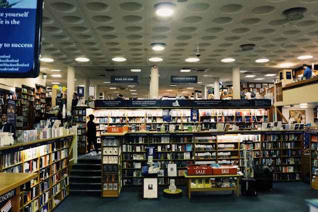 A spacious bookstore with multiple shelves filled with books, including a sale section in the center. The store features a modern design with a high ceiling and circular light fixtures. Several people are browsing the books, and signs such as 'PLEASE PAY HERE' and 'CUSTOMER SERVICES' are visible.