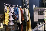 A selection of colorful costumes hangs on a clothes rack in front of a sign labeled 'HAIR & MAKEUP.' The costumes include a yellow ruffled dress, a silver fringe dress, and other vibrantly textured garments. There's a handwritten note about the close relationship between the makeup designers and actors alongside images of the team at work.