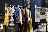 A selection of colorful costumes hangs on a clothes rack in front of a sign labeled 'HAIR & MAKEUP.' The costumes include a yellow ruffled dress, a silver fringe dress, and other vibrantly textured garments. There's a handwritten note about the close relationship between the makeup designers and actors alongside images of the team at work.