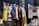A selection of colorful costumes hangs on a clothes rack in front of a sign labeled 'HAIR & MAKEUP.' The costumes include a yellow ruffled dress, a silver fringe dress, and other vibrantly textured garments. There's a handwritten note about the close relationship between the makeup designers and actors alongside images of the team at work.