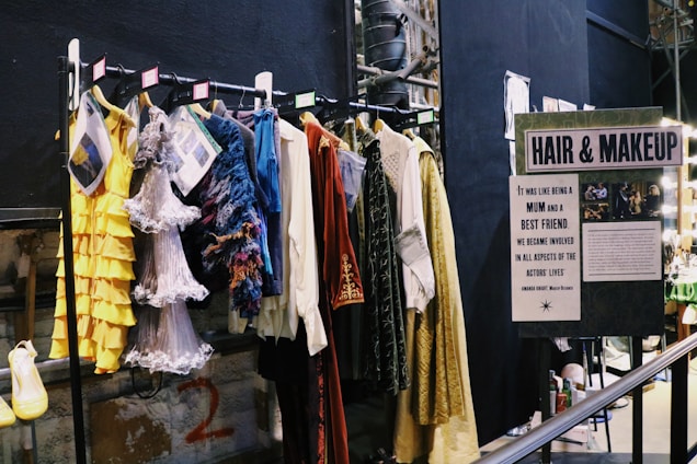 A selection of colorful costumes hangs on a clothes rack in front of a sign labeled 'HAIR & MAKEUP.' The costumes include a yellow ruffled dress, a silver fringe dress, and other vibrantly textured garments. There's a handwritten note about the close relationship between the makeup designers and actors alongside images of the team at work.