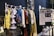 A selection of colorful costumes hangs on a clothes rack in front of a sign labeled 'HAIR & MAKEUP.' The costumes include a yellow ruffled dress, a silver fringe dress, and other vibrantly textured garments. There's a handwritten note about the close relationship between the makeup designers and actors alongside images of the team at work.
