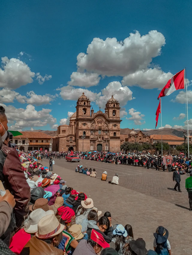 Crowds in traditional dress fill Cusco's Plaza de Armas before the twin-towered Compañía de Jesús church, Peruvian flags flying under a cloud-filled sky.
