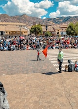 A crowded public square with a large group of people gathered, some sitting and others standing. A child holding a red flag is walking across a cobblestone area while a uniformed person, possibly a security officer, looks on. The background features colonial-style buildings with tiled roofs and a mountainous landscape under a partly cloudy sky.