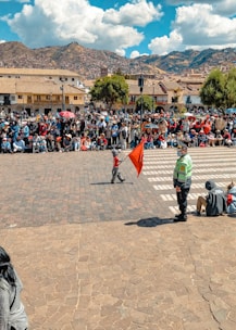 A crowded public square with a large group of people gathered, some sitting and others standing. A child holding a red flag is walking across a cobblestone area while a uniformed person, possibly a security officer, looks on. The background features colonial-style buildings with tiled roofs and a mountainous landscape under a partly cloudy sky.