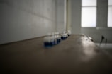 A series of small glass jars with blue liquid are lined up on a long, wooden surface in a dimly lit room. The image has a shallow depth of field, blurring the surrounding area and background, which includes large windows allowing diffuse light to enter.