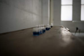 A series of small glass jars with blue liquid are lined up on a long, wooden surface in a dimly lit room. The image has a shallow depth of field, blurring the surrounding area and background, which includes large windows allowing diffuse light to enter.