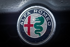 A close-up of the Alfa Romeo logo on a vehicle, covered in water droplets. The emblem features a red cross on the left and a green serpent on the right, encircled by a metallic border with the brand name in white on a blue background.