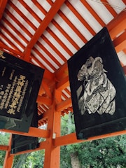 A detailed view of a traditional Japanese architectural structure featuring wooden beams painted in bright orange and white. There are two black plaques hanging, one displaying Japanese calligraphy and the other depicting an intricate artwork, possibly of a historical or religious figure. Lush green foliage is visible in the background.