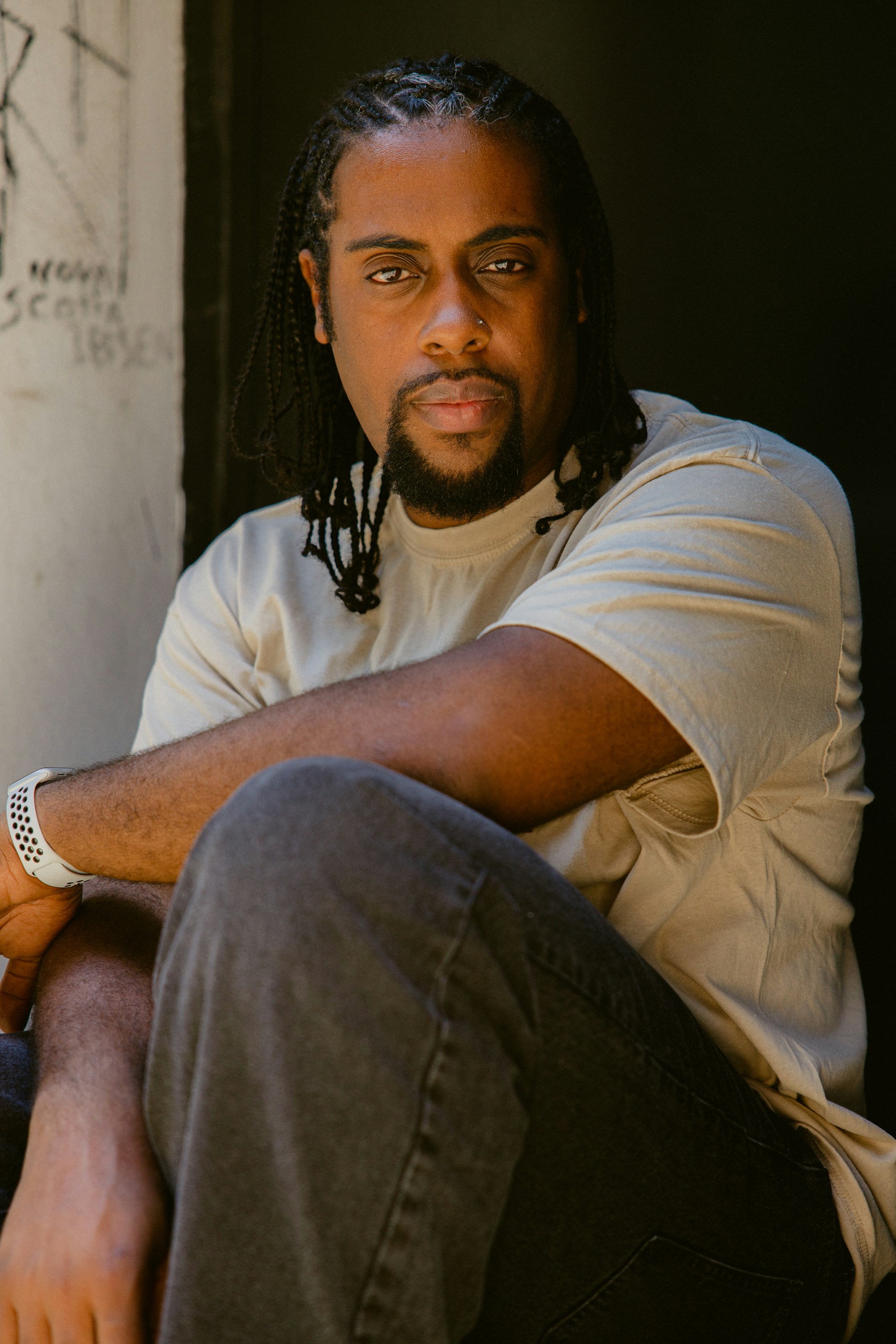 A man with braided hair and a beard sits with his arm resting on his knee. He wears a casual light-colored t-shirt and dark pants, with a white wristband on his right arm. The background is dimly lit, with some graffiti visible on a nearby wall.