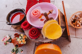 A variety of colorful objects are placed on an uneven surface. There's a pink basin with carved wax flowers, a black pot filled with red liquid, bundles of reddish-brown wax, and a stack of yellow wax discs. A ceramic pitcher with floral decorations and a basket containing various tools are also visible.