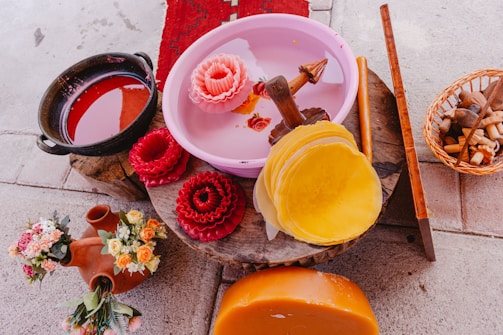 A variety of colorful objects are placed on an uneven surface. There's a pink basin with carved wax flowers, a black pot filled with red liquid, bundles of reddish-brown wax, and a stack of yellow wax discs. A ceramic pitcher with floral decorations and a basket containing various tools are also visible.
