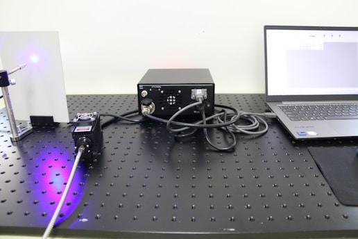 A laboratory setup includes a laser device emitting a blue light, a control box with cables, and a laptop displaying data or software. The components are arranged on a perforated optical table.