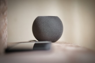 A compact, spherical smart speaker with a textured mesh exterior sits on a surface. In the foreground, part of a smartphone is visible, slightly out of focus. The background is softly blurred, giving the image a minimalistic and modern feel.