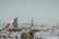 A panoramic view of a cityscape with the Eiffel Tower prominently in the background. In the foreground, a French flag is visible, flying atop a building. The landscape is densely packed with classic architecture and rooftops, with hints of greenery scattered throughout.