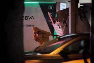 A person wearing a yellow raincoat appears to be carrying something on their back while crouching. There is a background featuring a sign with the word 'BBVA' and currency symbols. The setting seems urban, possibly near a bank or financial institution, with dim lighting and a partially visible car in the foreground.