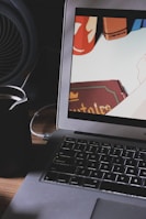 A laptop on a wooden desk displaying an animation scene on its screen. Beside the laptop is a black cup, possibly holding a beverage. The background includes a fan, and the screen shows colorful books or objects.
