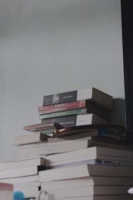 A stack of books is placed on a flat surface, with the books piled unevenly. Titles such as 'Emma' by Jane Austen and 'Moby Dick' by Herman Melville are visible, along with several Conan comic books. The surface also holds some small objects, possibly personal items like a bottle or container. The lighting is dim and casts soft shadows.