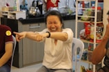 A person sitting on a chair is laughing joyfully, pointing with both hands towards something or someone. The background shows a kitchen area with various kitchenware items like a kettle and a shelf with snacks and other food items.