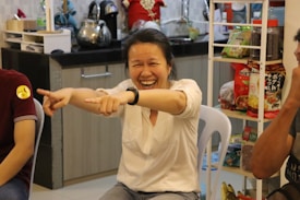 A person sitting on a chair is laughing joyfully, pointing with both hands towards something or someone. The background shows a kitchen area with various kitchenware items like a kettle and a shelf with snacks and other food items.