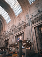 An interior setting of a grand architectural space with high vaulted ceilings and intricate stone carvings adorning the walls. The structure features large archways and a glass ceiling allowing natural light to illuminate the space. Below, there are signs for food establishments, including 'Pastaria' and 'Storie & Caffe'.