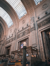 An interior setting of a grand architectural space with high vaulted ceilings and intricate stone carvings adorning the walls. The structure features large archways and a glass ceiling allowing natural light to illuminate the space. Below, there are signs for food establishments, including 'Pastaria' and 'Storie & Caffe'.