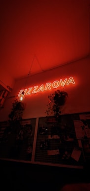 A warm red neon sign displaying the word 'PIZZA' hangs above a dimly lit wall. Decorative plants are mounted on the wall, with their leaves cascading downward. Below the sign, a wire grid features various papers, images, and notes attached to it, giving the space an eclectic and cozy vibe.