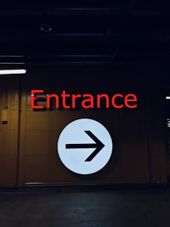 A dimly-lit interior space features a wall with a large illuminated white circle displaying a black right-pointing arrow. Above the circle is the word 'Entrance' in bold red letters.