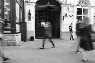 A black and white photograph of a street scene with blurred figures walking. A man stands still against a building in the background, looking at his phone. The building features arched windows and decorative elements. The street is paved, and there are brick and glass surfaces visible to the left.