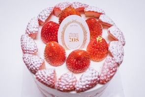 A beautifully decorated cake with fresh strawberries and powdered sugar on top. There is a decorative card in the center with golden lettering and a design that adds elegance to the presentation.