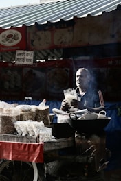 A street vendor is standing behind a cart filled with packets of puffed snacks. The vendor is holding a bag of snacks, surrounded by a variety of snack packages. The scene is partially shadowed, with sunlight streaming through parts of a corrugated metal roof above. Smoke is visible near the vendor, possibly from cooking or roasting.