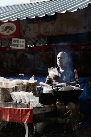 A street vendor is standing behind a cart filled with packets of puffed snacks. The vendor is holding a bag of snacks, surrounded by a variety of snack packages. The scene is partially shadowed, with sunlight streaming through parts of a corrugated metal roof above. Smoke is visible near the vendor, possibly from cooking or roasting.