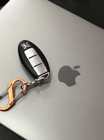 A car key fob with a metallic orange carabiner attached rests on a silver laptop with an Apple logo.