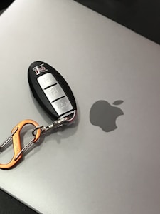 A car key fob with a metallic orange carabiner attached rests on a silver laptop with an Apple logo.