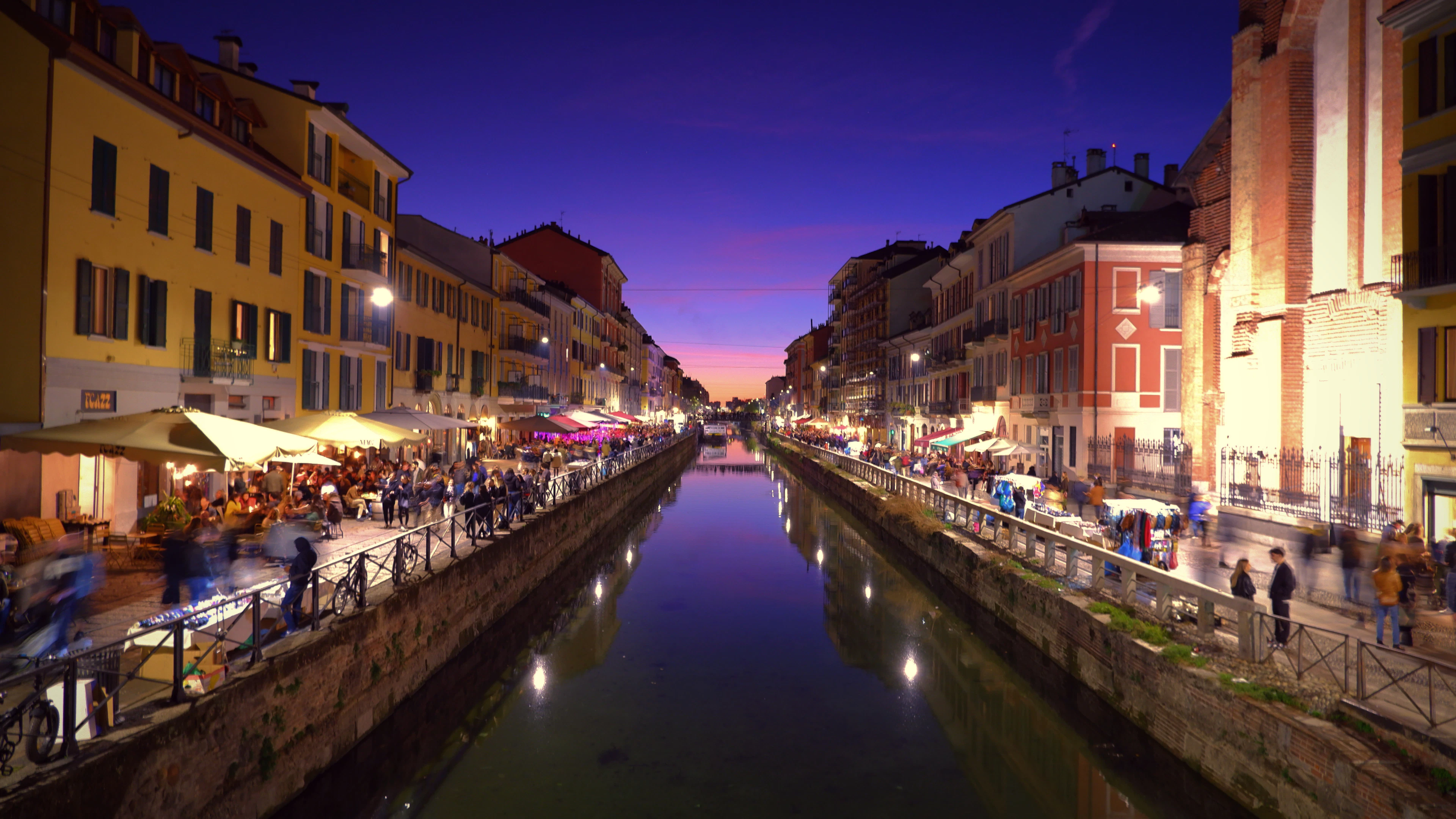 Milan's Naviglio Grande at dusk, lit restaurant terraces lining both stone quays beneath a violet sky and mirrored in the still canal water.