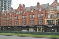 A row of historic red-brick buildings with detailed architectural features, including arched windows and decorative brickwork. The ground floor houses several businesses with black signs, such as Quarter Horse Coffee Roasters and a barbershop. A modern construction project is visible in the background, contrasting with the older architecture. In front of the buildings, there is a low hedge dividing the sidewalk from the street.