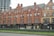 A row of historic red-brick buildings with detailed architectural features, including arched windows and decorative brickwork. The ground floor houses several businesses with black signs, such as Quarter Horse Coffee Roasters and a barbershop. A modern construction project is visible in the background, contrasting with the older architecture. In front of the buildings, there is a low hedge dividing the sidewalk from the street.