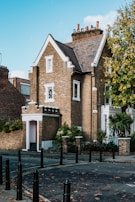 A charming brick house with a pitched roof and several chimneys, surrounded by greenery and set against a bright blue sky. The building features detailed masonry work and white-trimmed windows. It's located on a quiet street with black bollards lining the sidewalk.
