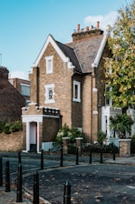 A charming brick house with a pitched roof and several chimneys, surrounded by greenery and set against a bright blue sky. The building features detailed masonry work and white-trimmed windows. It's located on a quiet street with black bollards lining the sidewalk.
